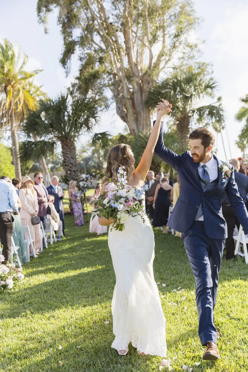 Kara in her wedding dress with her bouquet, hand raised with Kevin’s, walking back up a grass aisle between guests under palms, 14 March 2026