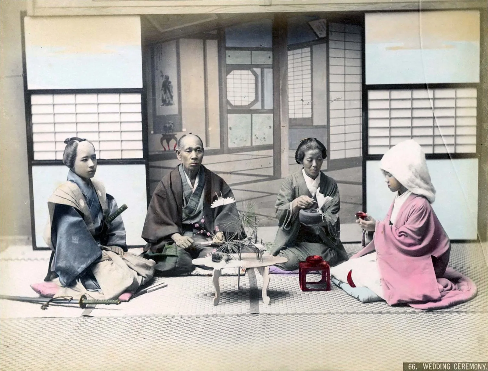 A hand-coloured studio photograph: a young groom with swords, an older couple, and a bride in a white hood kneel around a low table holding a pine