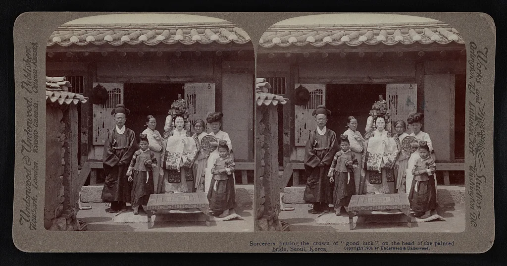 A stereograph: a bride in embroidered robes and a tall crown stands on a veranda with her family and children