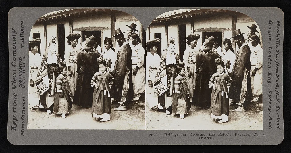 A stereograph: a bridegroom in a black horsehair hat bows before the bride’s family outside a house