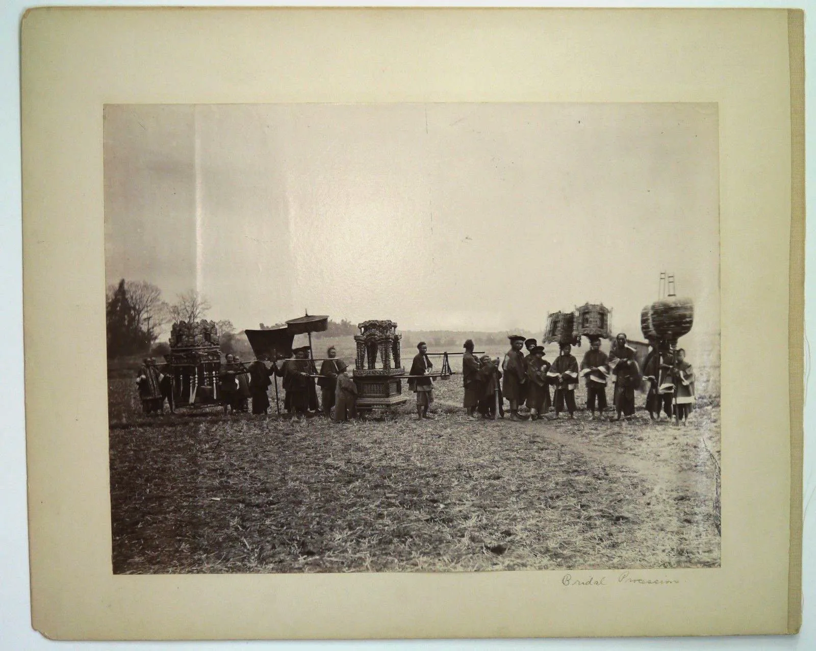 A mounted albumen print of a bridal procession in open country: bearers with a covered sedan chair, ornamental pavilions on poles, and a parasol