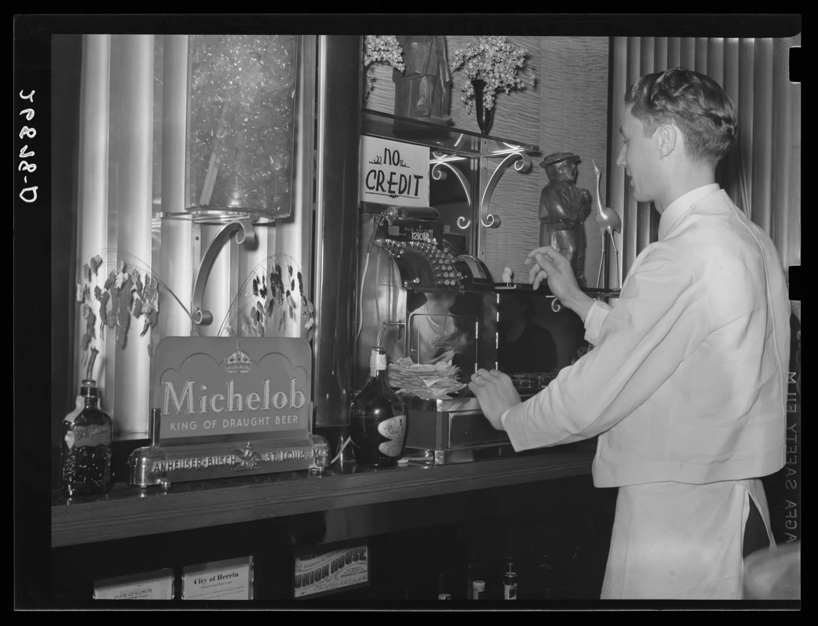 A bartender in a white jacket and bow tie behind a long wooden bar in the Rainbow Room of the Ly-Mar Hotel, Herrin, Illinois, 1939